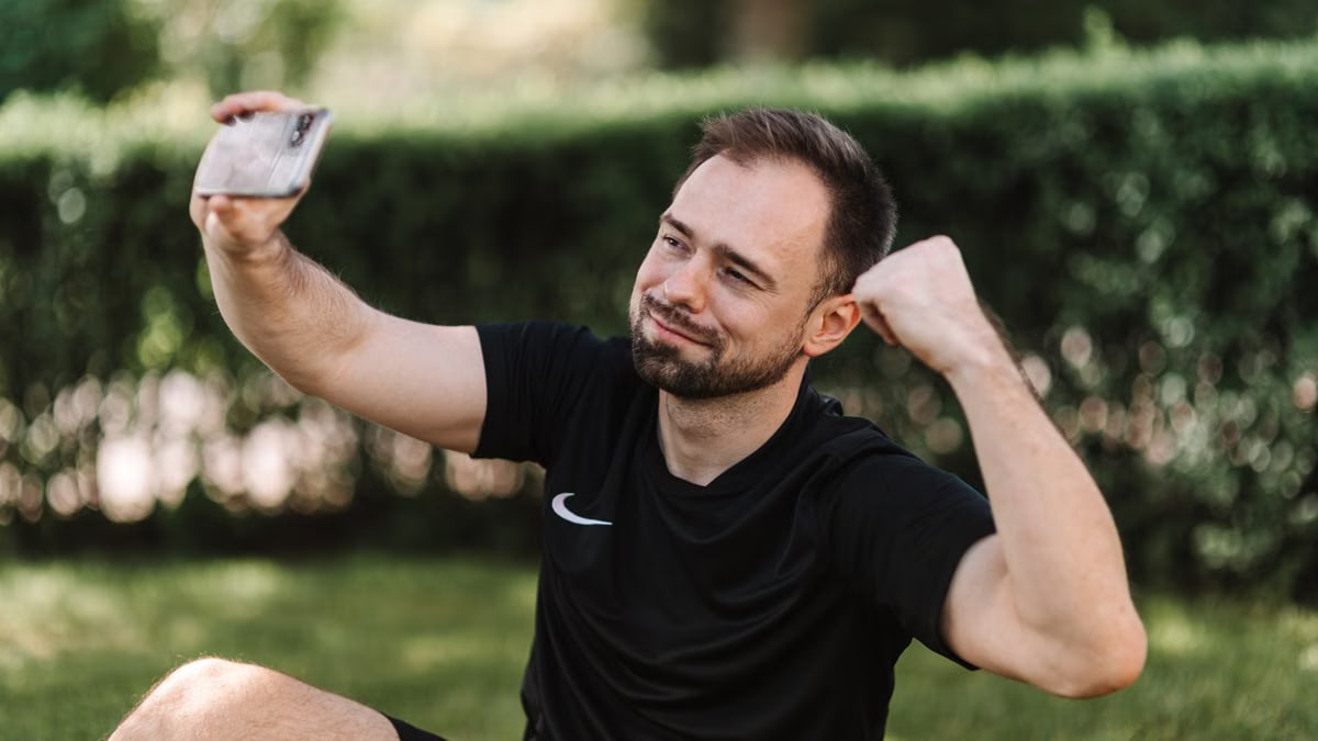 Smiling man in black shirt taking a selfie outdoors, flexing his arm.